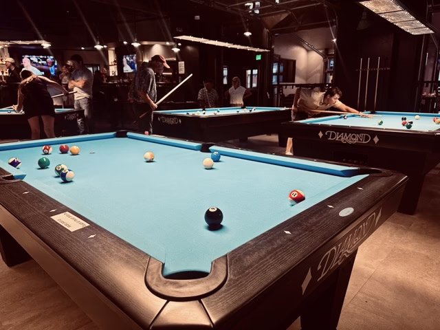Four friends enjoy a lively game at coopersmiths-billiards—two play while two watch and smile; one holds a drink. The casual bar setting features warm lighting, creating the perfect atmosphere for fun and relaxation.