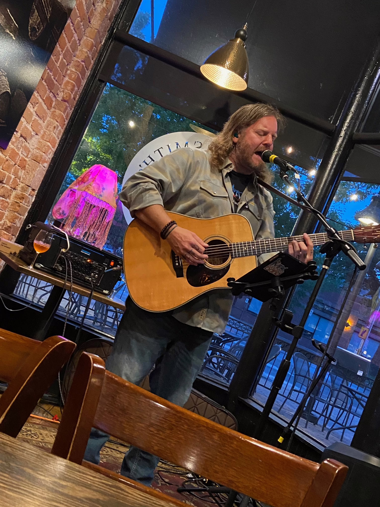 IMG_8822 IMG_8822 shows a long-haired, bearded man playing acoustic guitar and singing into a microphone at a cozy indoor venue, with a glass of beer on the table beside him and a colorful lamp in the background.