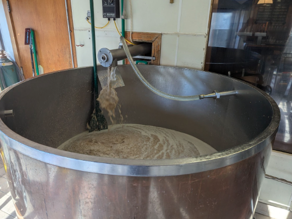 A large OMBABrewing metal brewing tank, partially filled with liquid and grain, is shown. A transparent hose is dispensing more grain mixture into the tank. Brewing equipment and a broom are visible in the background.