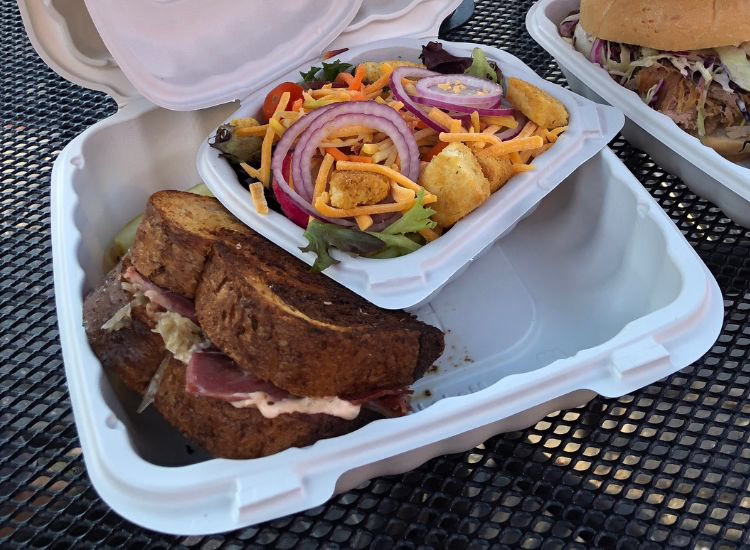 A takeout container holds a grilled sandwich with meat and cheese, and a small salad topped with sliced red onions, shredded cheese, croutons, and assorted greens. Another sandwich is partially visible in the background.