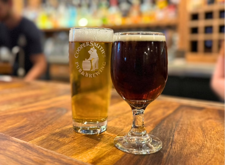beer-billiards Two glasses of beer-billiards rest on a wooden table: one is a tall, light lager with a frothy head and “Coopersmith’s Pub & Brewing” logo, the other is a dark amber pour in a goblet-style glass.