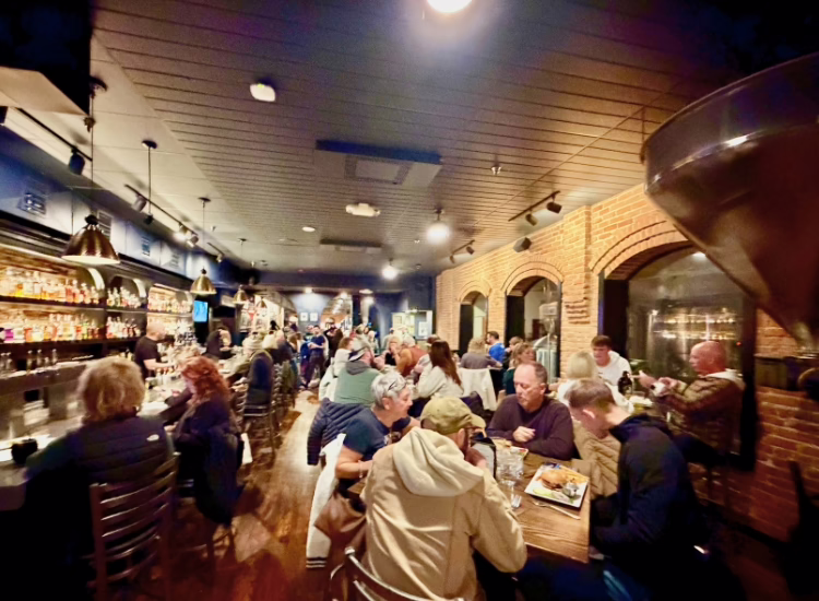The brewpub-dining scene features people enjoying meals at tables and the bar. Warm lights, exposed brick walls, and large windows create a cozy, bustling atmosphere filled with lively conversation.