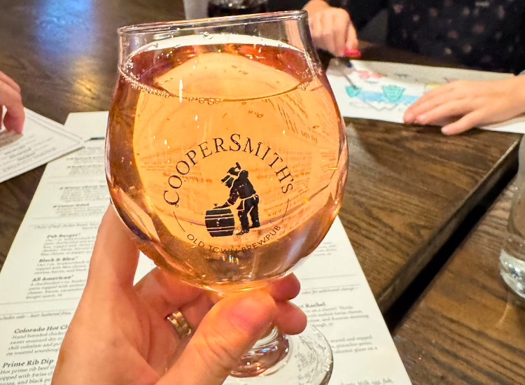 A hand holds a glass of cocktails with the "Coopersmith's Old Town Brewpub" logo. Menus rest on the wooden table, while two people sit in the background—one is coloring on paper.