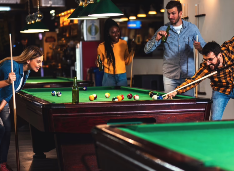 Four friends enjoy a lively game at coopersmiths-billiards—two play while two watch and smile; one holds a drink. The casual bar setting features warm lighting, creating the perfect atmosphere for fun and relaxation.