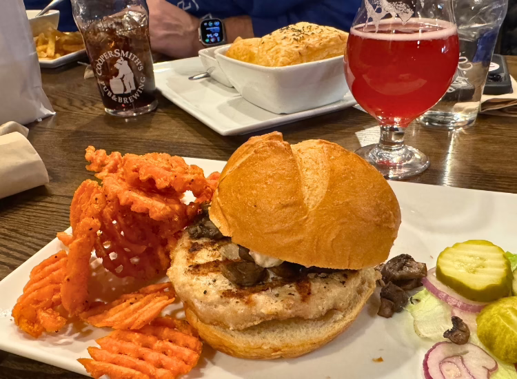 A coopersmiths-turkeyburger on a bun with mushrooms, served with waffle fries, pickles, and sliced onions on a white plate. A red drink, water, and another meal sit on the wooden table.