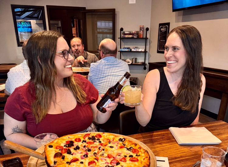 Two women sit at a restaurant table, smiling and toasting with drinks—one holding a beer, the other a cocktail. A large pizza is in front of them, and other diners are visible in the background.